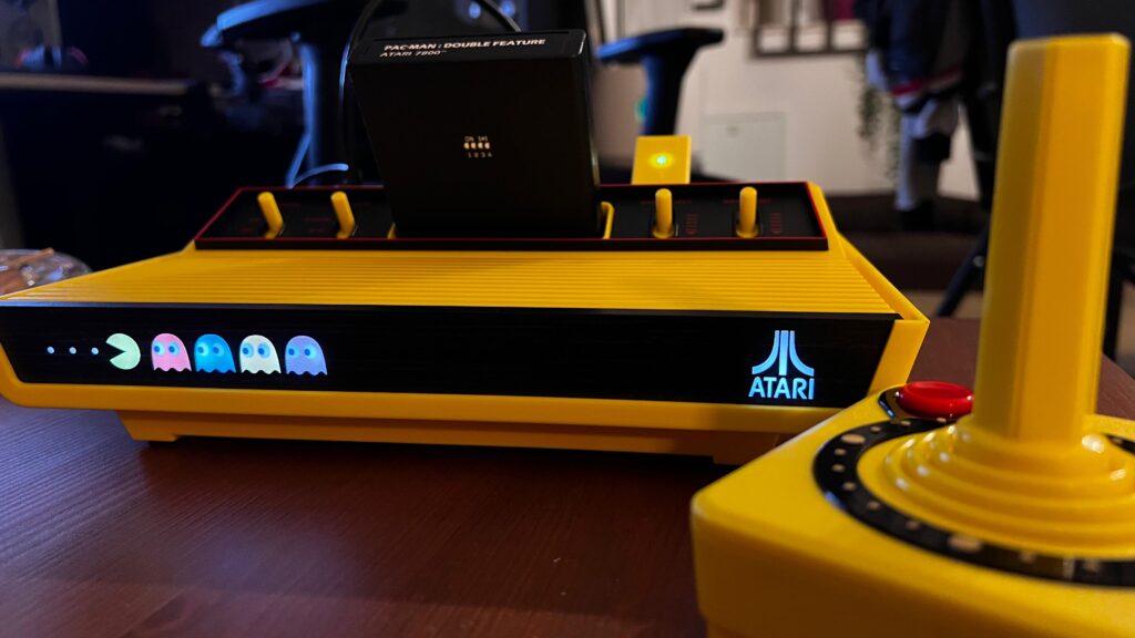 yellow Atari 2600 Plus Pac-Man edition powered on with ATARI sign lighting on the right of the console, blinky, pinky, inky and dinky on the left chasing pacman and on the right to the console the directional controller with pacman details on it